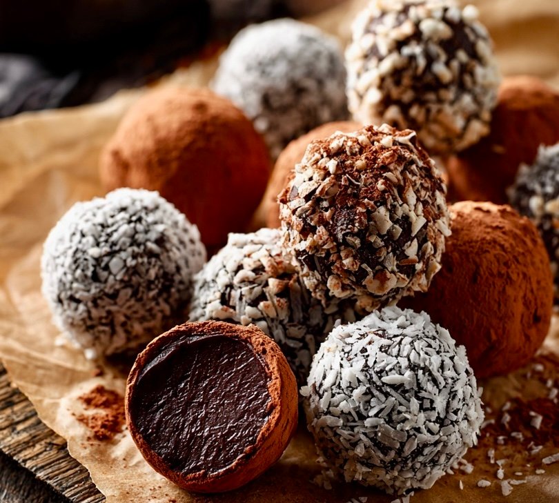 Bright overhead view of assorted chocolate ganache truffles on crinkled parchment paper — cocoa‑dusted, powdered sugar‑coated, and glossy sprinkle‑covered truffles surrounded by bowls of cocoa powder, melted chocolate, powdered sugar, and chocolate sprinkles on a rustic wooden table.