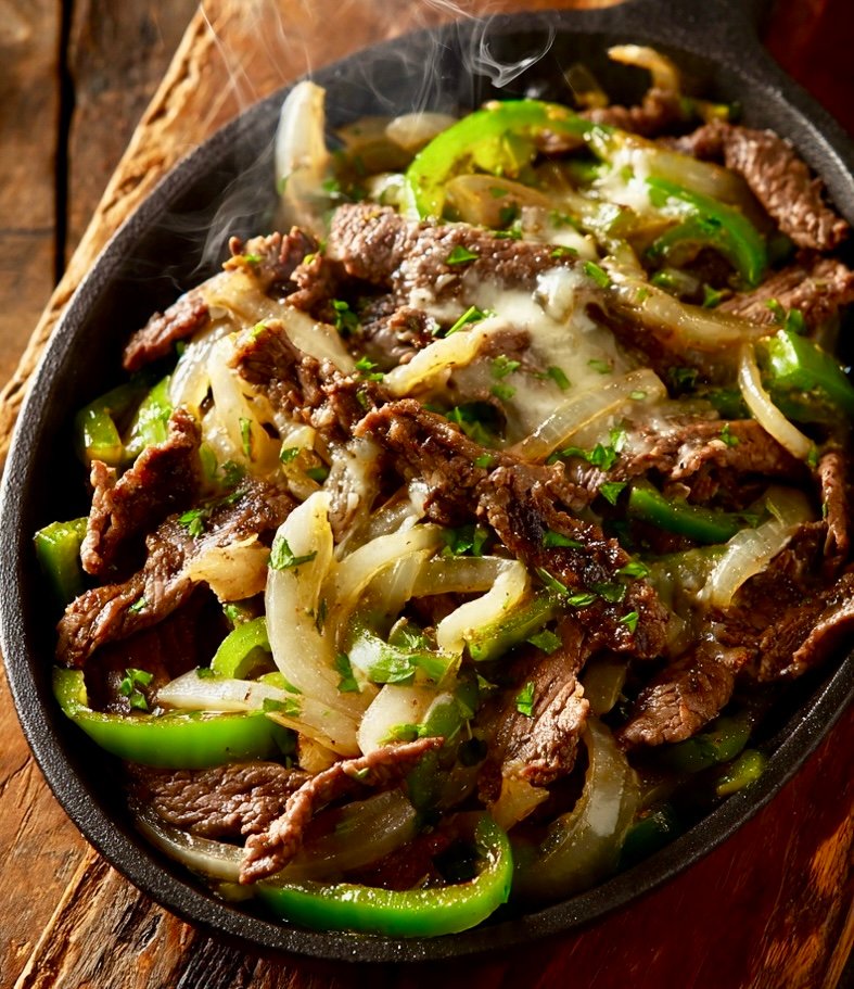 30 minute Philly Cheesesteak. Overhead view of an electric skillet filled with sizzling Philly cheesesteak — thinly sliced beef, sautéed green bell peppers and onions, topped with melted provolone cheese, steam rising, on a rustic wooden table.