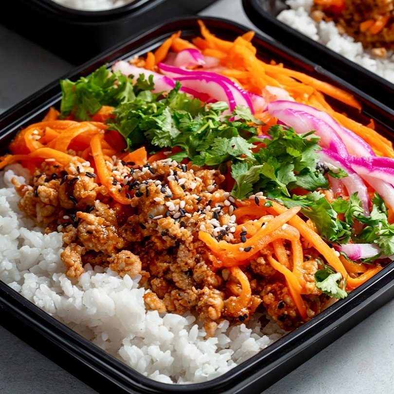 Banh Mi Chicken Bowls . A black meal prep container filled with fluffy white rice, savory ground chicken, bright orange carrots, fresh green cilantro, and bright purple pickled red onions