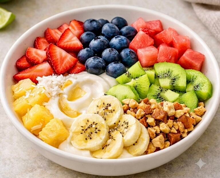 A wide white bowl filled with a colorful, no-cook summer yogurt bowl. The center contains a swirl of creamy white yogurt drizzled with honey, surrounded by neat rows of sliced strawberries, blueberries, watermelon cubes, chopped kiwi, sliced bananas, chopped pineapple, and a handful of chopped nuts.